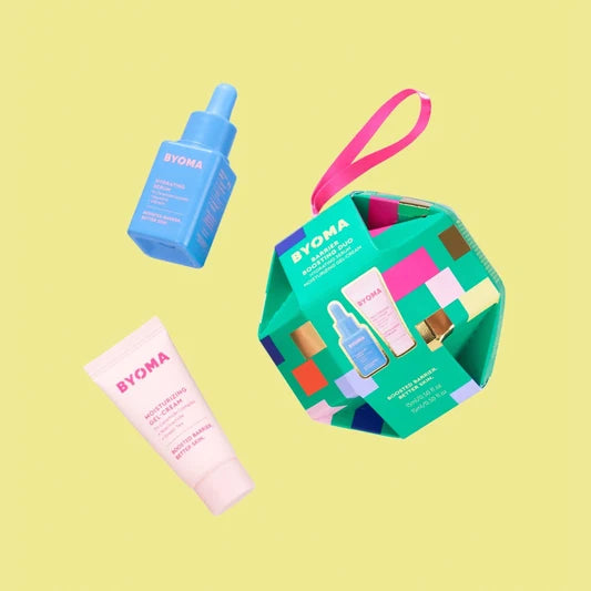 Un coffret cadeau en forme d'ornement vert multicolore intitulé « BYOMA Barrier Boosting Duo », avec un tube rose de gel-crème hydratant et un flacon bleu de sérum hydratant. Les produits flottent sur un fond jaune.